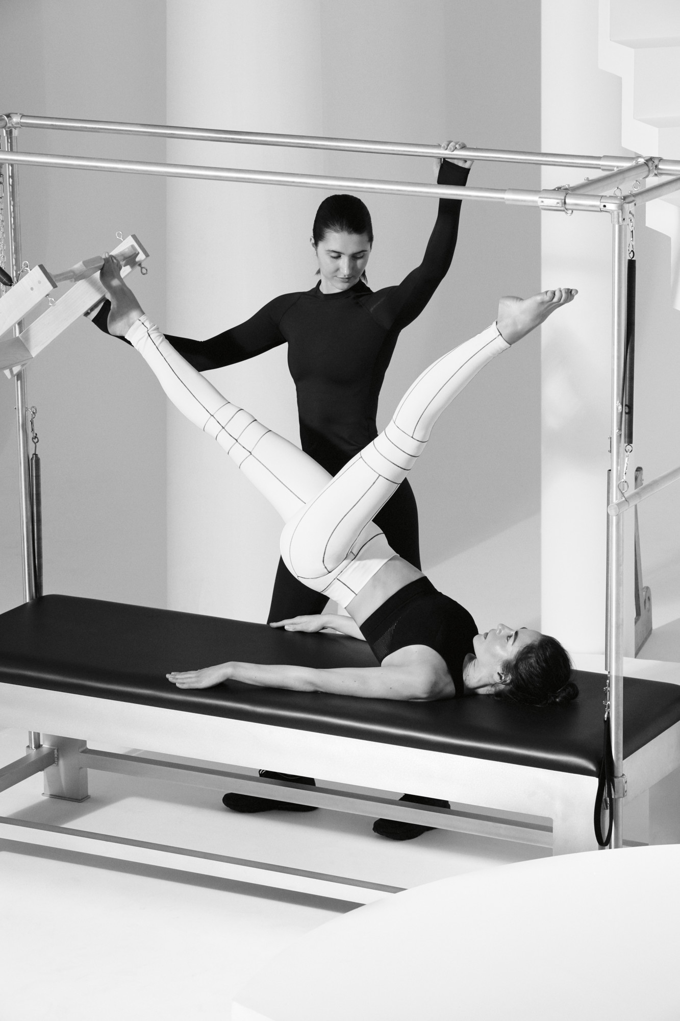<black and white image of woman doing pilates on cadillac reformer with pilates instructor