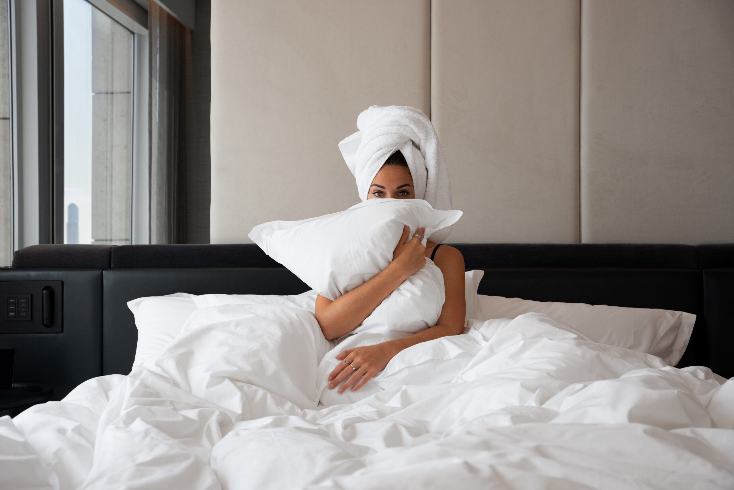 Woman in hotel bed holding a pillow with her arms and a towel in her hair